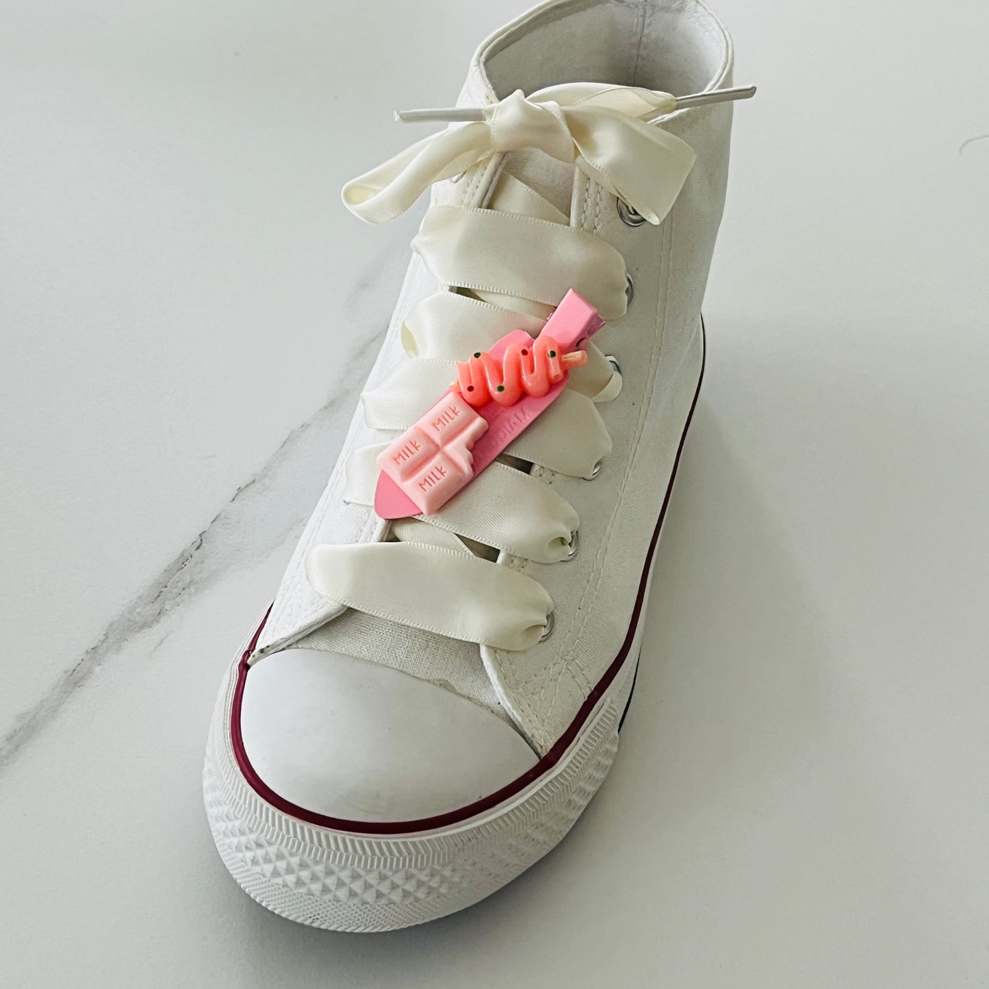 une converse avec un lacet écru en satin et une pince kawaii rose avec une tablette milk rose et une sucette collées sur la barrette